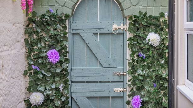 Une porte en bois rustique, dotée d’un loquet en métal, et encadrée d’une luxuriante végétation de lierre vert et de fleurs violettes.