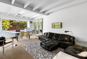 Uma sala de estar moderna com um sofá de couro preto, uma área de jantar com mesa e cadeiras, e uma grande janela com vista para o verde.