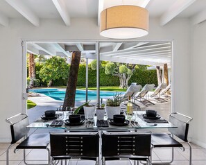Uma sala de jantar moderna com mesa e cadeiras de vidro, vista para uma piscina e área de estar ao ar livre com guarda-sóis.