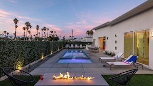 A modern outdoor pool area with lounge chairs, a fire feature, and a view of palm trees. - Picture Perfect Retreat – Modern Hollywood Glam (Palm Springs)