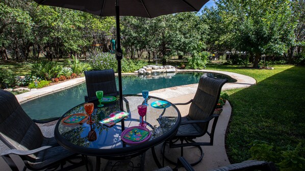 A round glass table with four chairs and a large umbrella, surrounded by a swimming pool and lush greenery. - Dripping Springs area Guesthouse Bungalow (Austin)