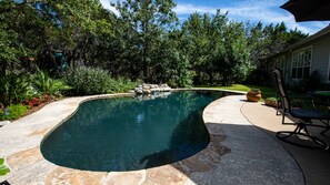 A swimming pool with a curved edge, surrounded by a stone deck and lush greenery. - Dripping Springs area Guesthouse Bungalow (Austin)