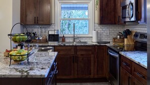 A kitchen with wooden cabinets, a stainless steel microwave, a double sink, and a window with a view of trees. - The Little River Farmhouse – Your Smoky Mountain Riverside Retreat (Townsend)