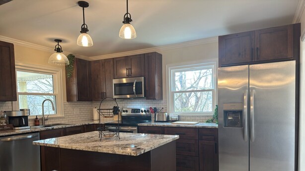 A modern kitchen with dark wooden cabinets, stainless steel appliances, a granite island with bar stools, and pendant lighting. - The Little River Farmhouse – Your Smoky Mountain Riverside Retreat (Townsend)