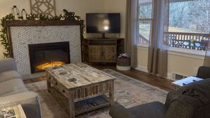 A living room with a fireplace, a television, and a view of the outdoors. - The Little River Farmhouse – Your Smoky Mountain Riverside Retreat (Townsend)