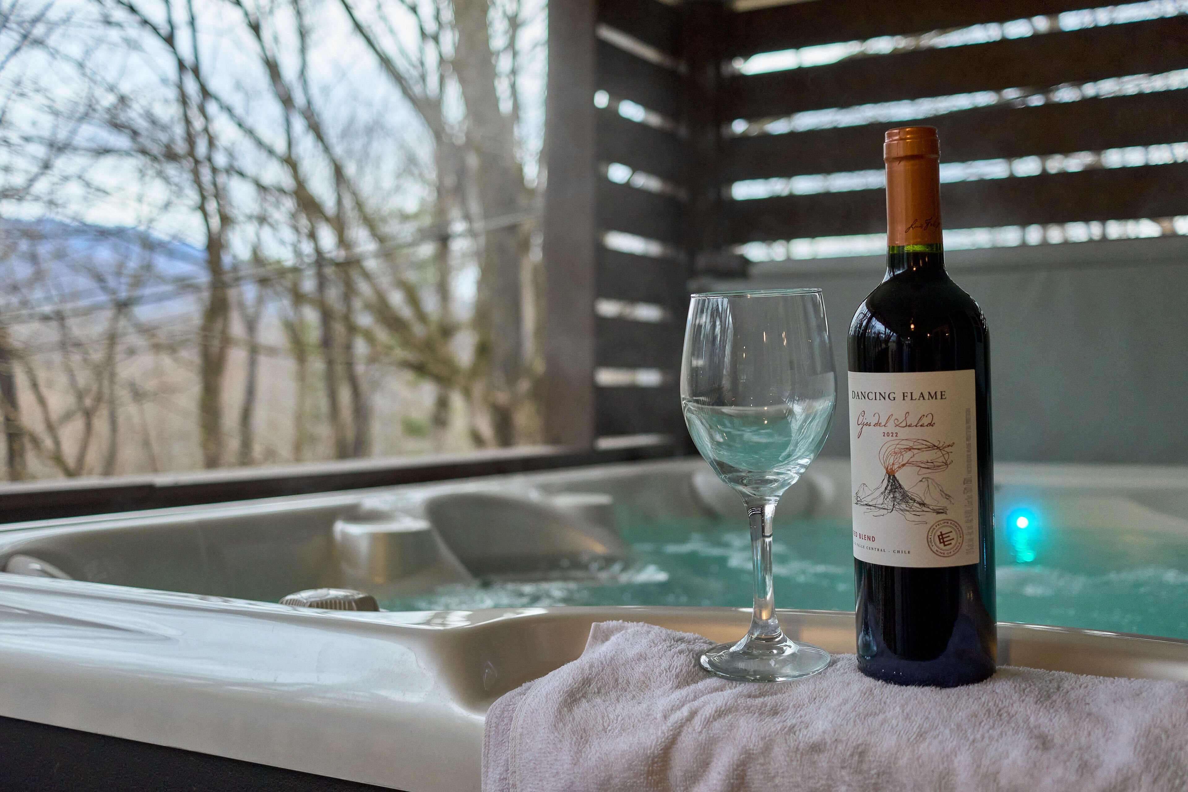 Uma garrafa de vinho Dancing Flame e um copo sobre uma toalha, perto de uma banheira de hidromassagem com vista para árvores e montanhas.