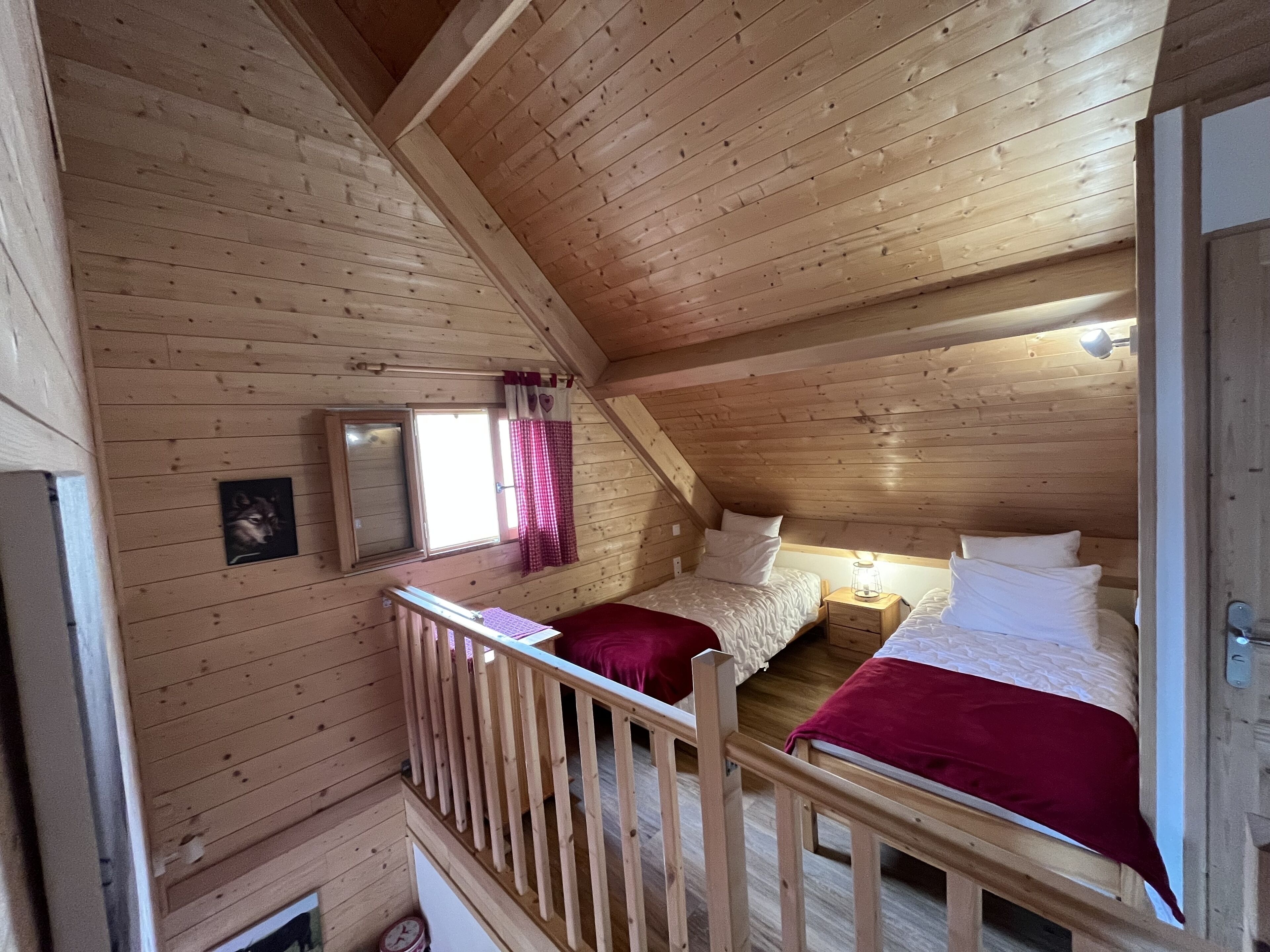 Ett mysigt sovrum på vinden med träväggar och ett sluttande tak. Det finns två sängar med rött och vitt sänglinne, ett litet fönster med en rutig gardin och en inramad tavla på väggen.
