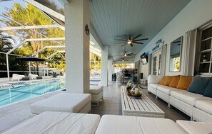 Un espacio exterior techado con piscina, mobiliario blanco, ventiladores de techo y vistas a la piscina.