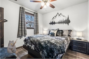 Un dormitorio con cama, mesita de noche, lámpara, ventana con cortinas, ventilador de techo y un adhesivo mural con montañas y árboles.