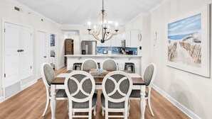 A dining area with a wooden table and white chairs, a chandelier, and a kitchen in the background.