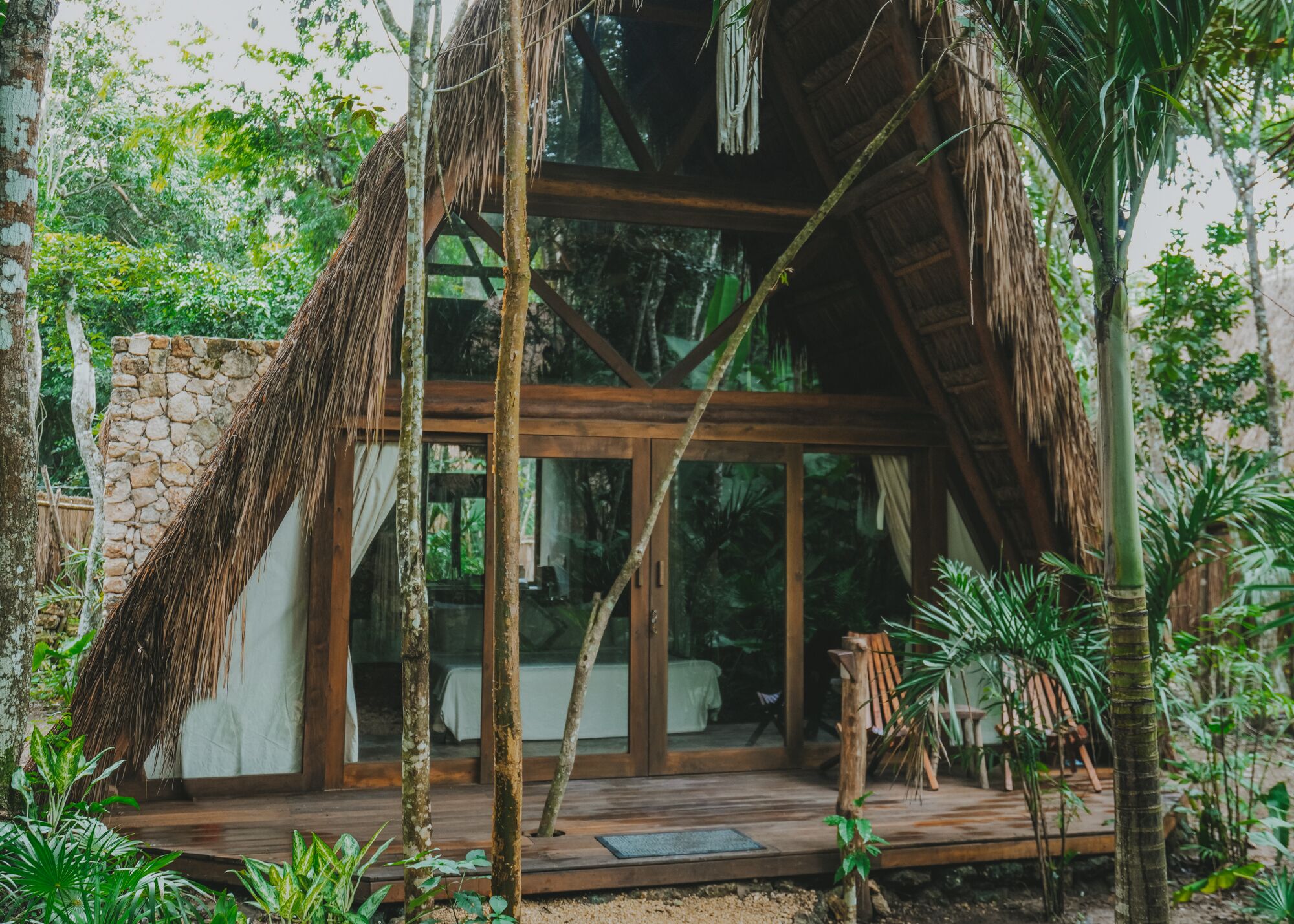 Una estructura con techo de paja, pared de piedra, terraza de madera y puertas de cristal que dan a una habitación.