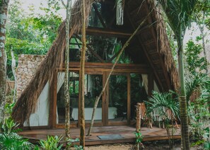 A thatched-roof structure with a stone wall, wooden deck, and glass doors leading to a room. - Naya Bacalar Hotel - Adults Only (Bacalar)