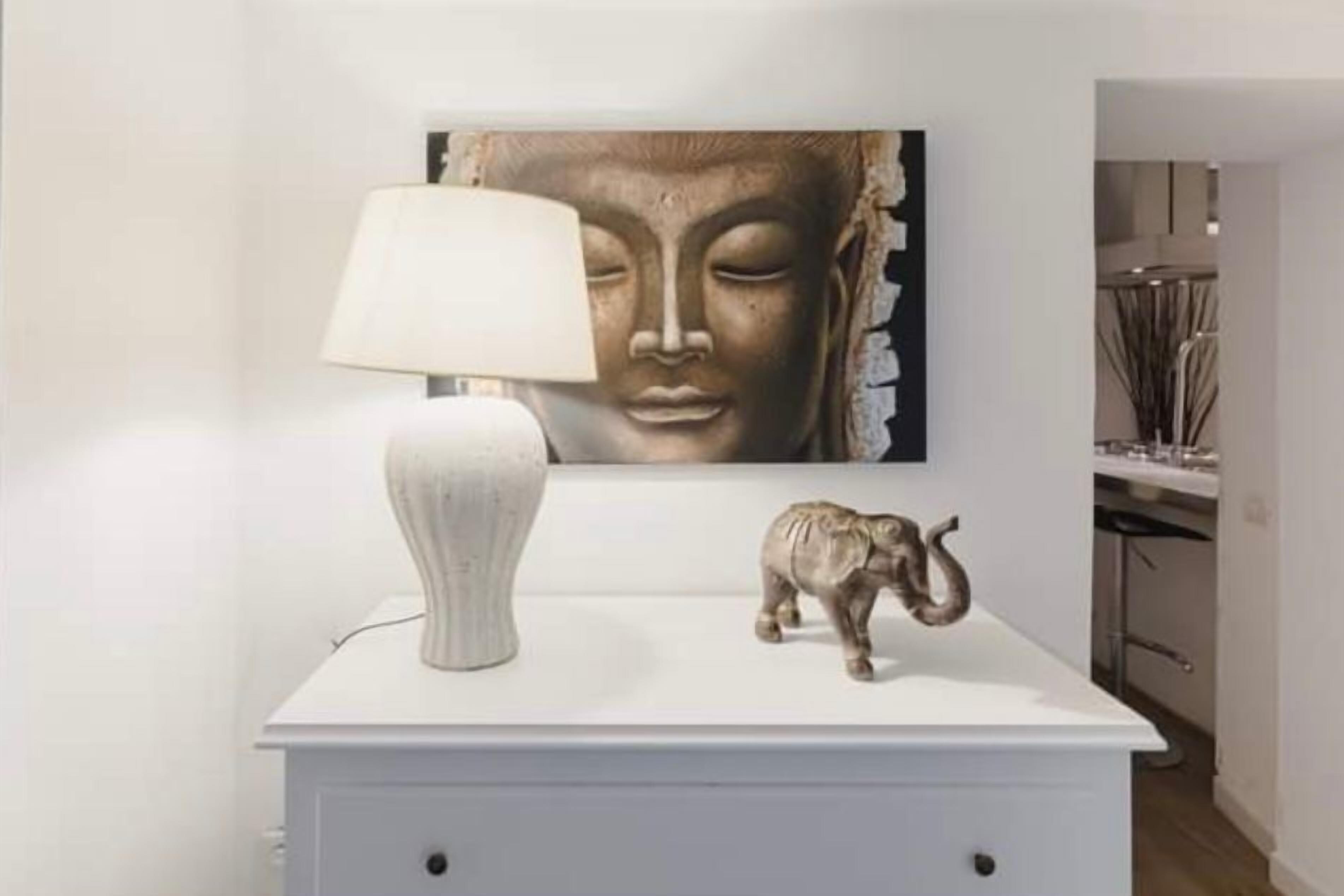 Et hvidt natbord med en lampe og en lille elefantfigur. Et stort Buddha-maleri hænger over bordet.