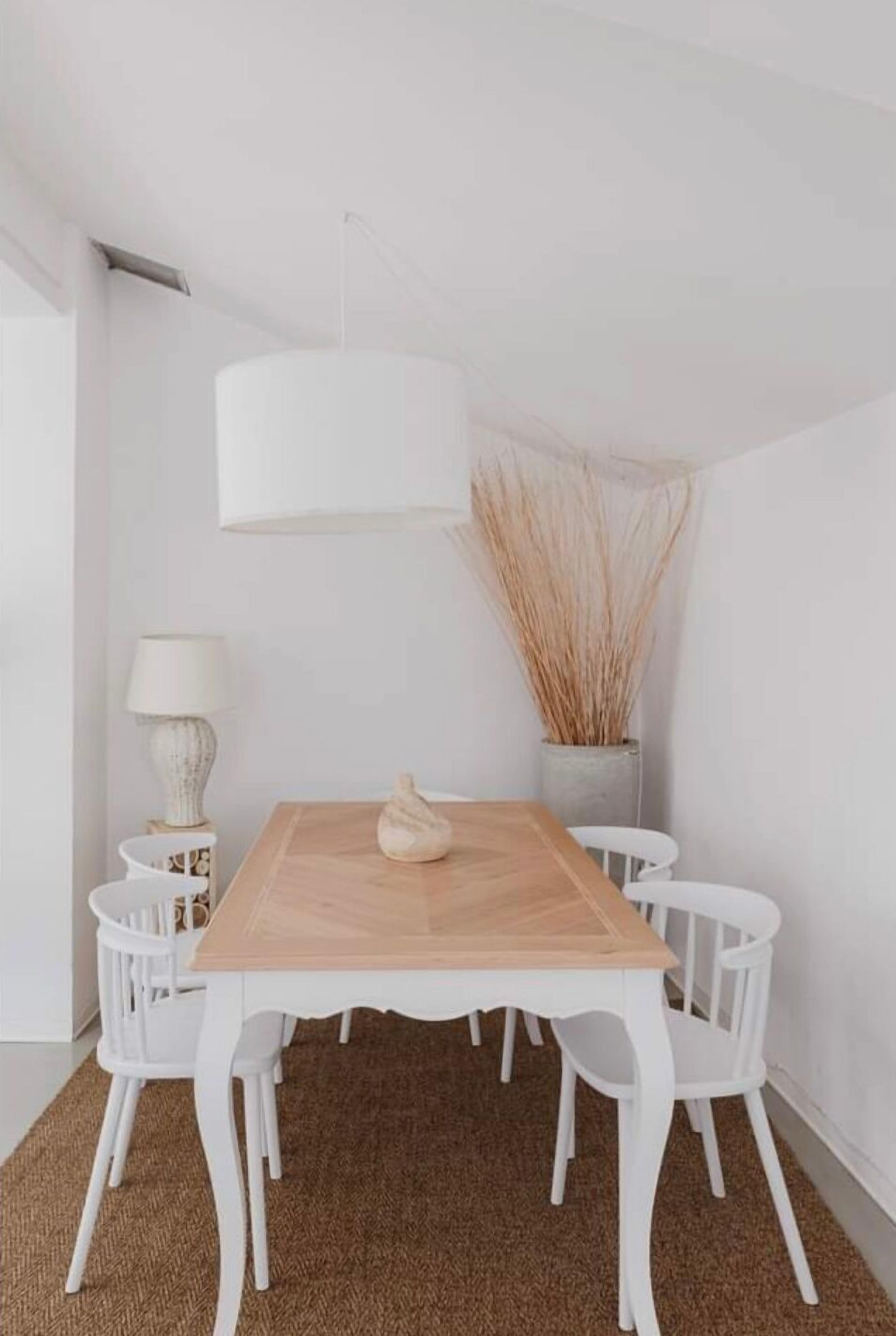 Ein Essbereich mit Holztisch, weißen Stühlen, einer weißen Lampe und einer Vase mit Trockenblumen.