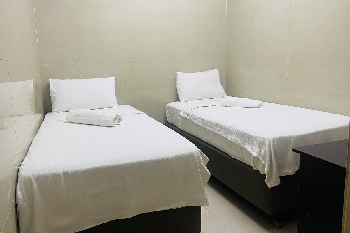 A room with two single beds, each with a white mattress and pillow, and a neatly folded white towel on one bed.
