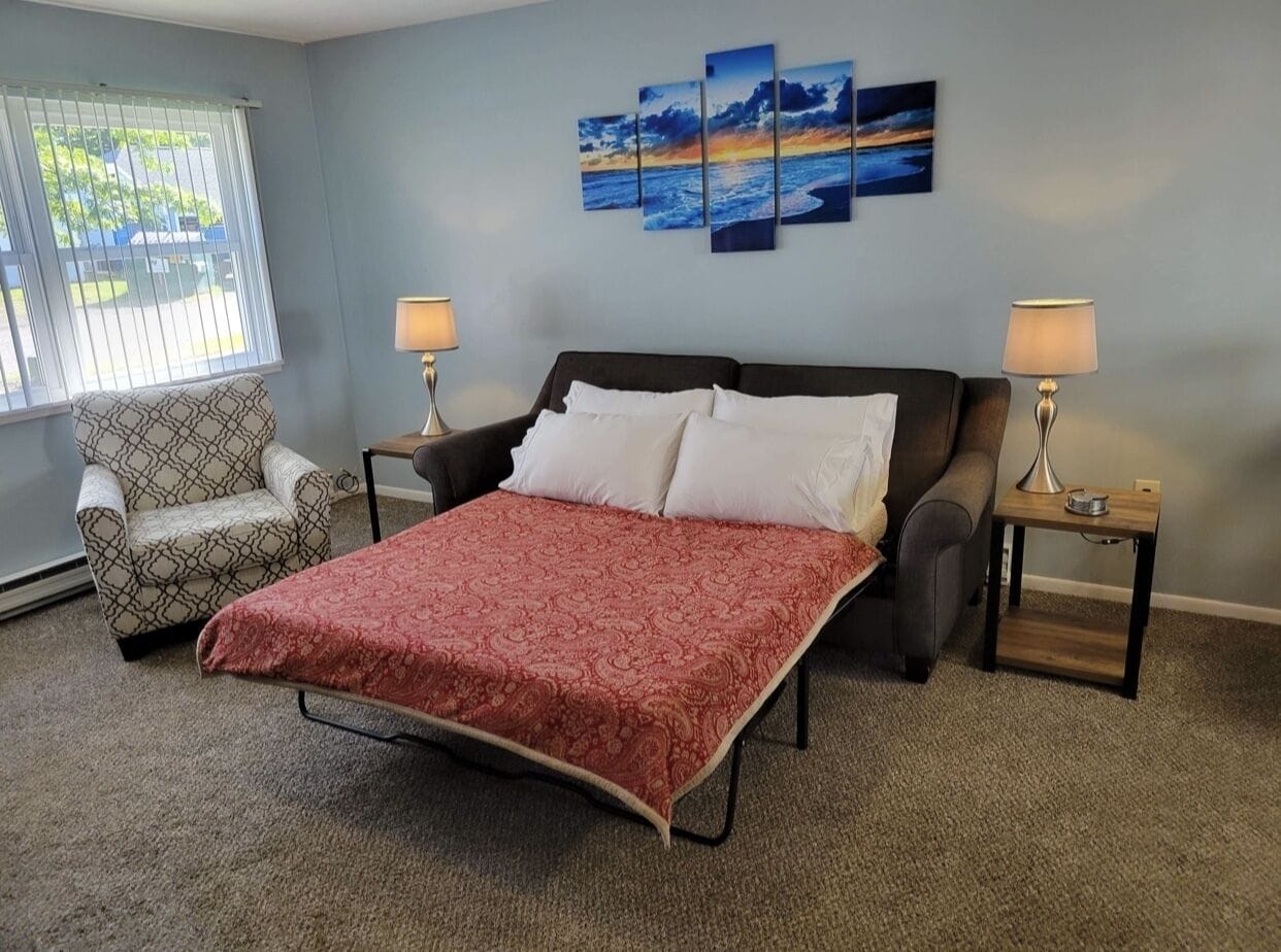 Quarto com cama, duas mesinhas de cabeceira com luminárias, uma poltrona estampada e uma parede com três quadros.