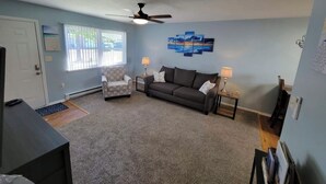A living room with a grey carpet, a grey sofa, a patterned armchair, a ceiling fan, and a wall with a blue artwork. - Tis’ Time Hideaway. Just a few blocks from beautiful Lake Erie!  (Conneaut)
