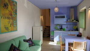 En kompakt opholdsstue med en grøn sofa, et spisebord med stole, et tekøkken med et fjernsyn og en trappe til et niveau ovenpå.