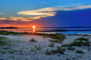 Une plage de sable avec une végétation clairsemée, un coucher de soleil en arrière-plan et une étendue d’eau calme.