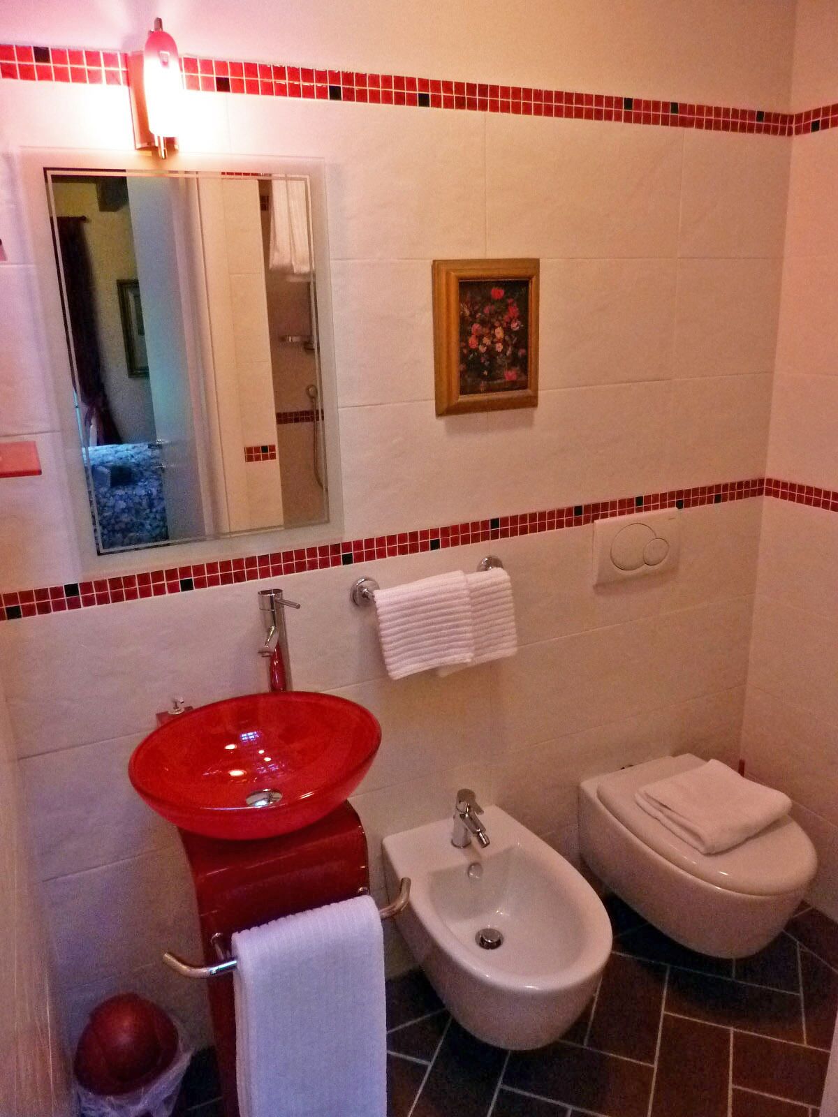 A bathroom with a red sink, a white toilet, and a bidet. There is a mirror, a towel rack, and a small framed picture on the wall.