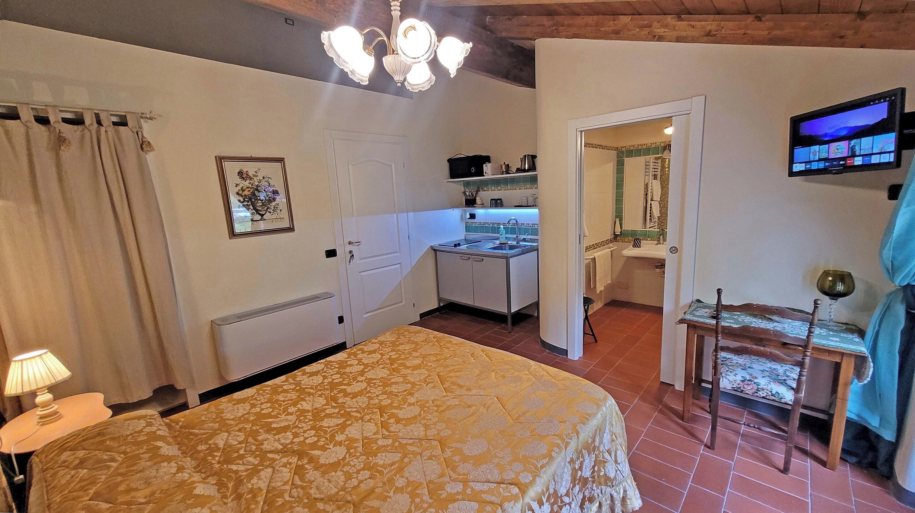A bedroom with a bed, a nightstand, a lamp, a ceiling light fixture, a door, and a view into a kitchen and bathroom.