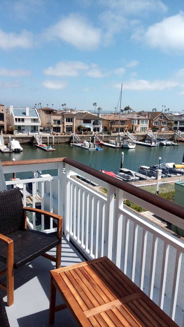 Un balcon avec un banc en bois et une chaise en osier, donnant sur une marina où l’on peut admirer des bateaux et des maisons.
