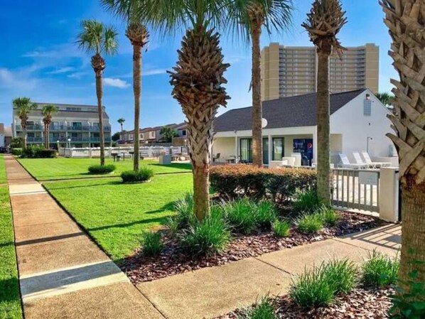 A hotel with a well-maintained lawn, palm trees, and a white building with a balcony. - Endless Summer E-12 Sleeps 4 - Just steps to beach! (Panama City Beach)