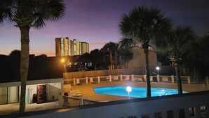 Vista nocturna de un área de piscina de hotel, con piscina, sillones de descanso y palmeras.