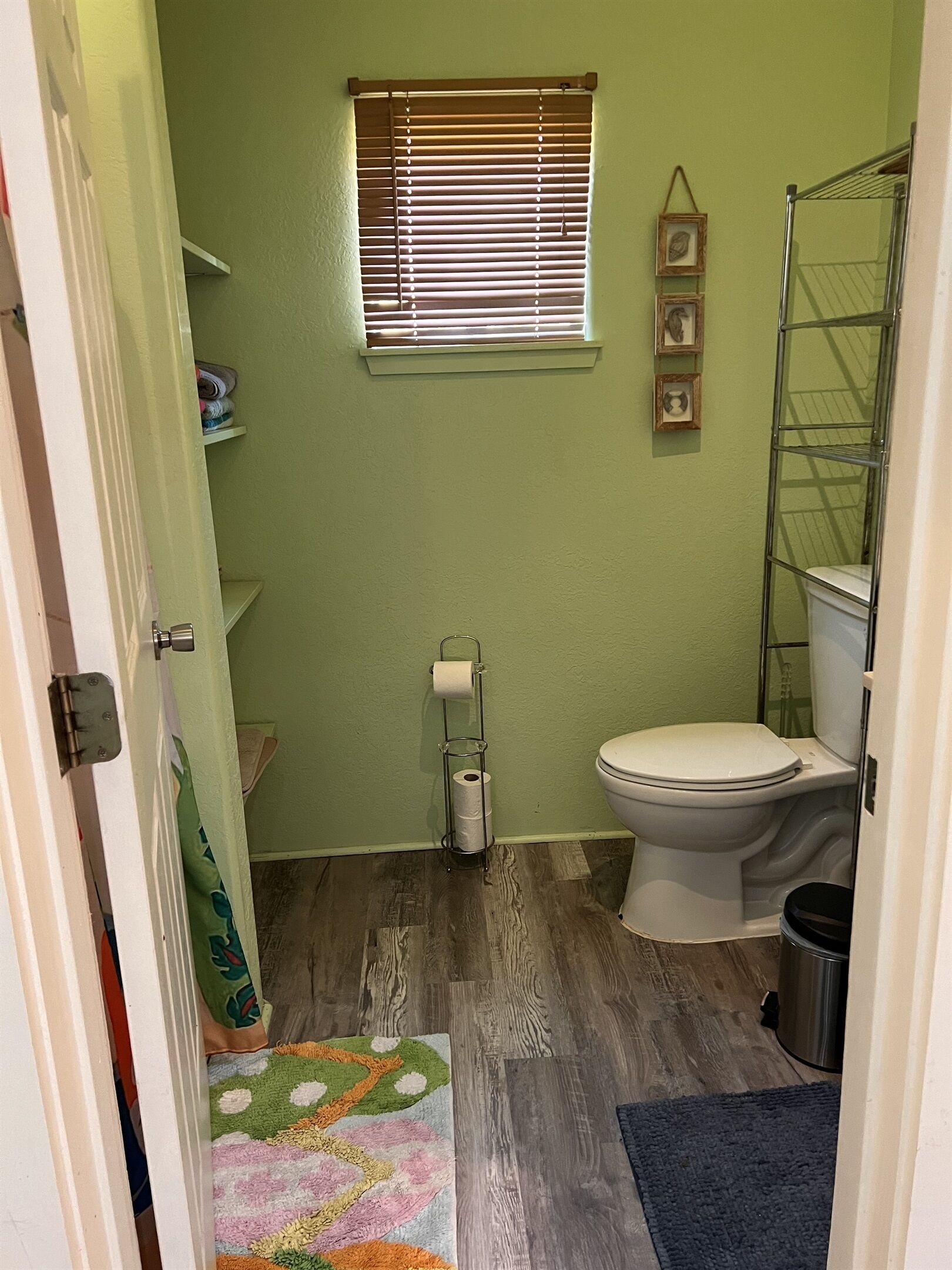 A small bathroom with a toilet, a window with blinds, a shower, and a metal rack.