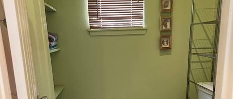 A small bathroom with a toilet, a window with blinds, a shower, and a metal rack.