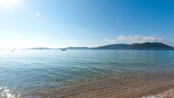 A clear beach with calm waters, distant mountains, and a few boats.