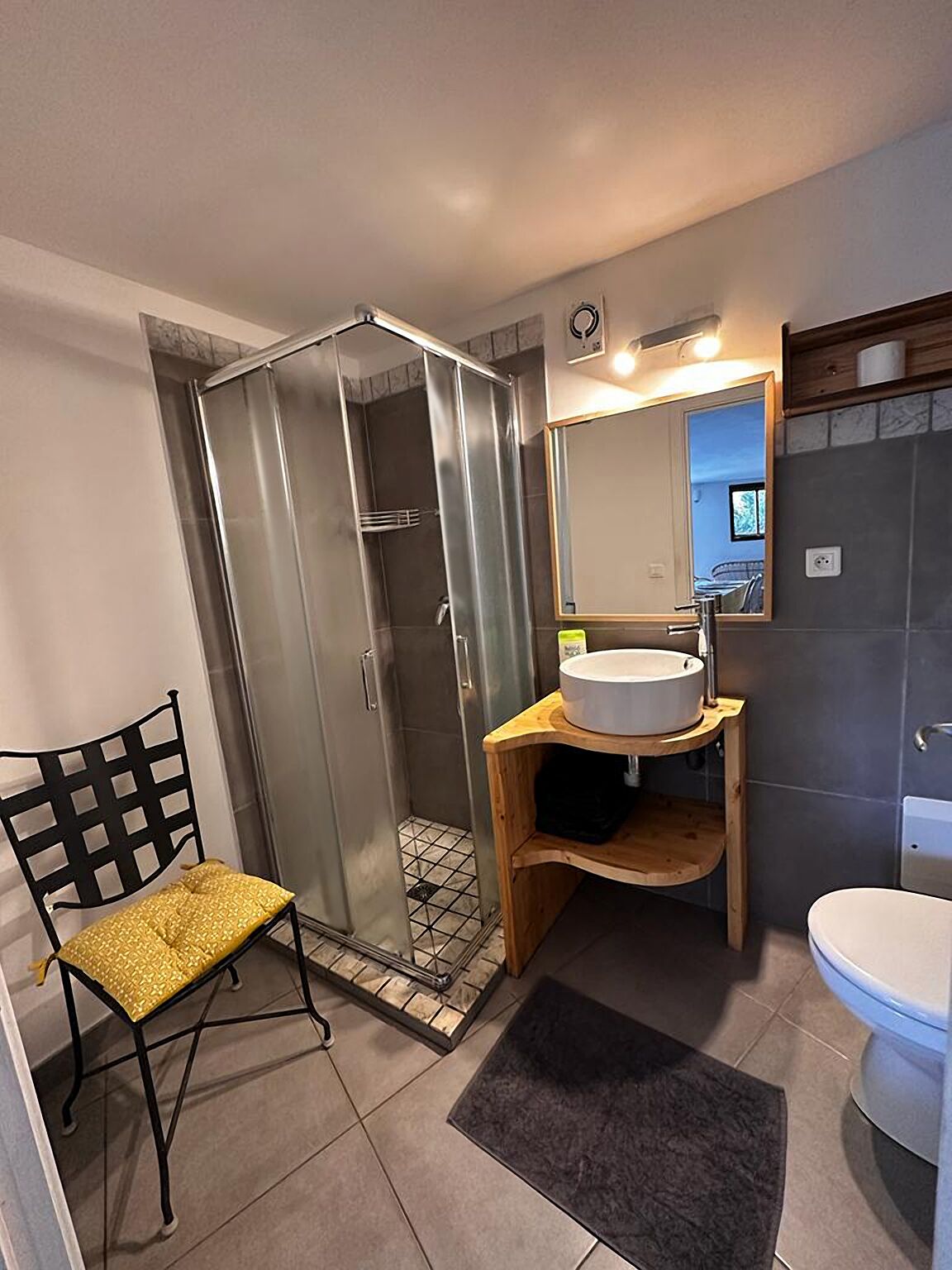 A bathroom with a glass-enclosed shower, a wooden vanity with a sink, a chair, and a toilet.