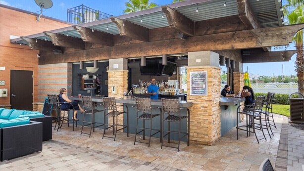 An outdoor bar area with a stone and metal structure, bar stools, and a seating area with a sofa.