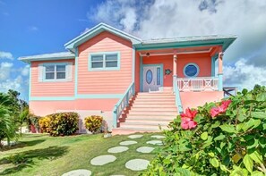 A two-story house with a pink and blue exterior, a front porch, and a small garden with stepping stones leading to the door.
