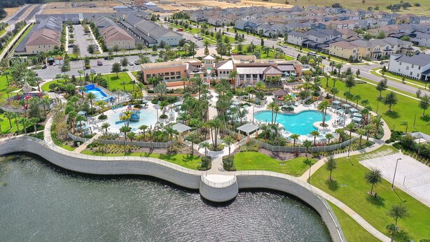 An aerial view of a residential area with a large swimming pool surrounded by houses, a bridge over water, and a road leading to a parking area.
