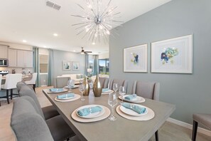 Un comedor moderno con una mesa larga dispuesta para seis personas, una cocina al fondo y dos cuadros en la pared.