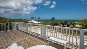 Une terrasse en bois avec une balustrade blanche, un banc blanc et une vue sur une plage bordée de palmiers, sous un ciel dégagé.