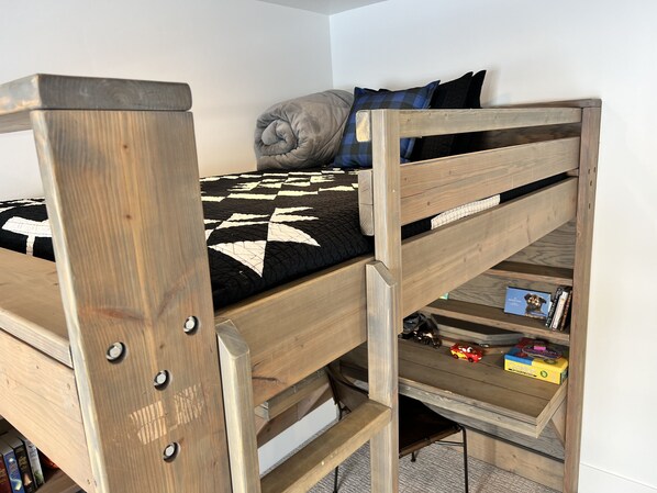 Una cama litera con estructura de madera y colcha blanco y negro. Hay un almohadón y una manta en la cama. Un estante debajo de la cama contiene libros y un coche de juguete.