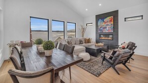 A spacious living room with a large wooden dining table, comfortable seating, a fireplace, and large windows offering a view of the landscape.