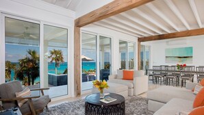 A spacious living area with a large sliding glass door leading to a balcony with a view of the ocean. There is a round coffee table with a vase of flowers, a sofa, and a dining area with chairs. - Sandy Hill Beach House 6 Bedroom Home (Governor's Harbour)