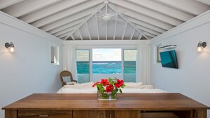 A bedroom with a wooden bed, a view of the ocean, a ceiling fan, and a TV mounted on the wall. - Sandy Hill Beach House 6 Bedroom Home (Governor's Harbour)