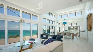 A spacious living area with a large window overlooking the ocean, a dining area with a kitchen, and a ceiling fan. - Dream House 3 Bedroom Home (North Palmetto Point)