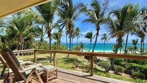 Une terrasse en bois avec deux chaises longues donnant sur une plage bordée de palmiers et sous un ciel bleu dégagé.