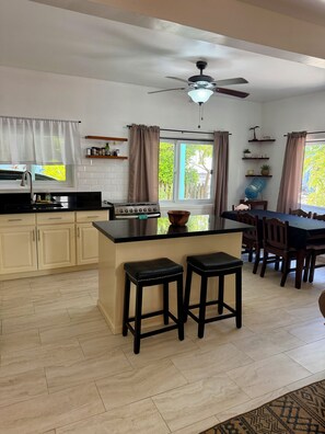 A kitchen with a central island, bar stools, a dining table with chairs, a ceiling fan, and a window with curtains. - On the beach, equipped with backup generator (West Bay)