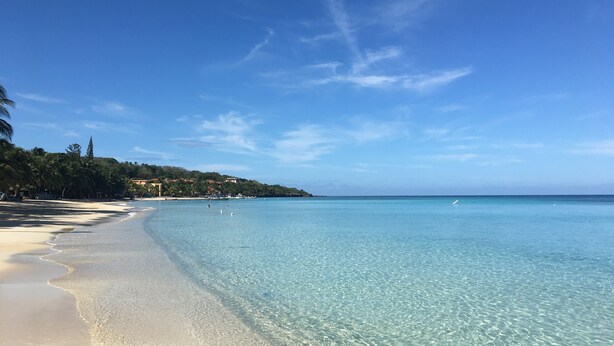Ein Strand mit klarem, türkisblauem Wasser, Sandküste und einem bewaldeten Hügel im Hintergrund.
