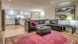 Un salon moderne avec un canapé en cuir, un pouf et un tapis à motifs rouges. On aperçoit une cuisine en arrière-plan, avec des meubles blancs et un coin repas.