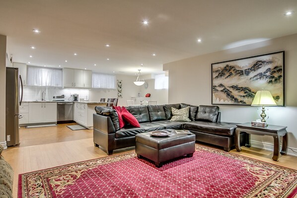 A modern living room with a leather sofa, ottoman, and a red patterned rug. There is a kitchen in the background with white cabinets and a dining area.