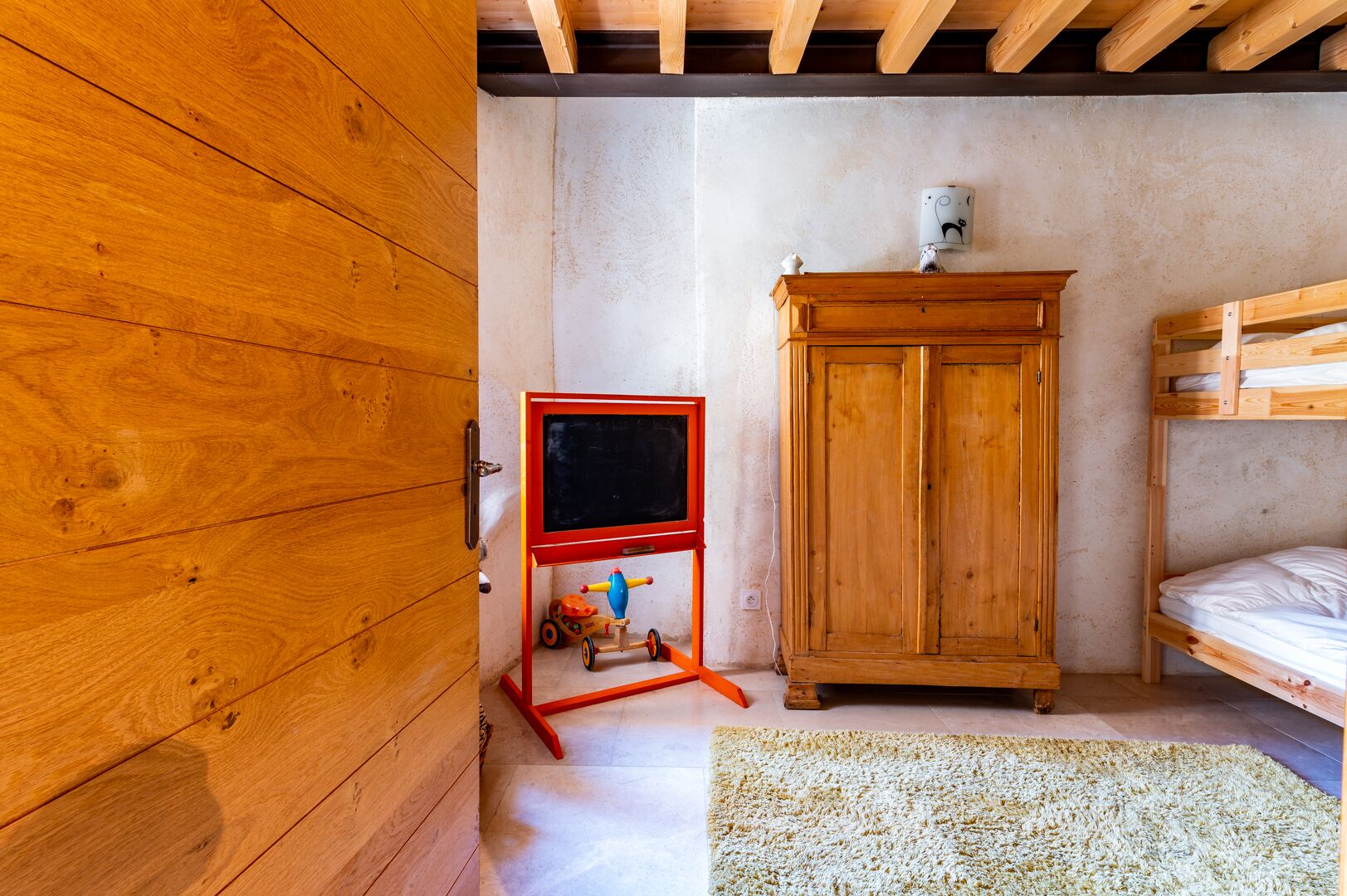 Una stanza con una porta in legno, un giocattolo rosso e blu su un piedistallo, un armadietto di legno e un letto a castello.