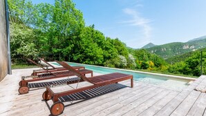 Eine Holzterrasse mit Liegestühlen und einem Pool, umgeben von üppiger Vegetation und Bergen.