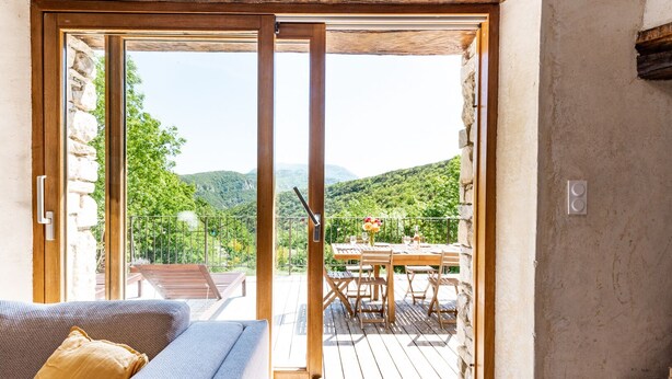 Ein Zimmer mit Holzbalkendecke, einer Schiebeglastür, die auf eine Terrasse mit Tisch und Stühlen führt, und einer Steinwand.